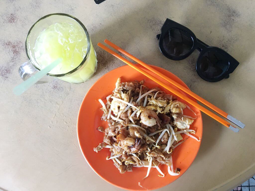 shrimp stir fry dish on orange plate with orange chopsticks. green drink to the left and black round-ish sunglasses to the right - where to eat in penang