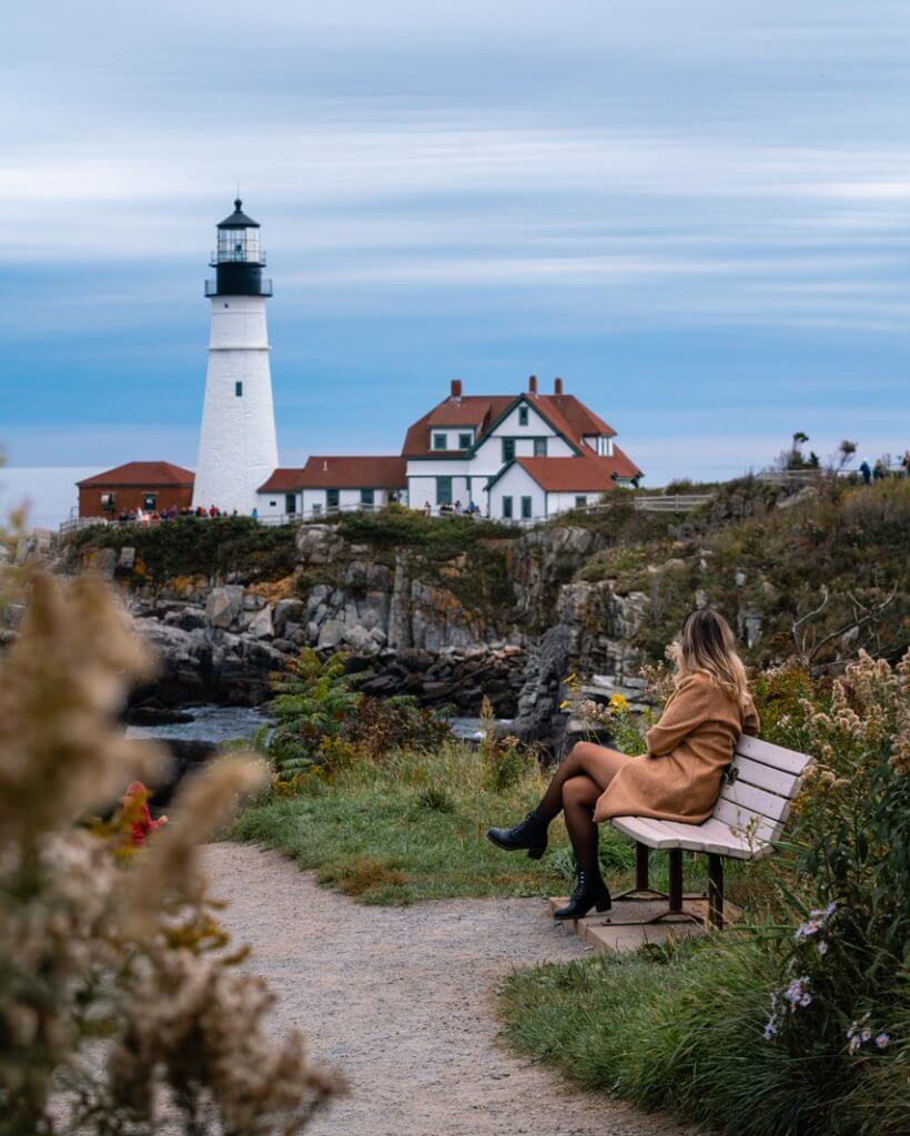 Megan sitting on a bench in Fort Williams Park taking in the view of Portland Head Lighthouse in Portland Maine