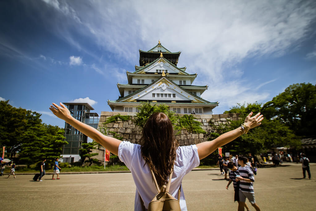 girl with long hair with arms up in front of 3-tiered Osaka Castle in Japan