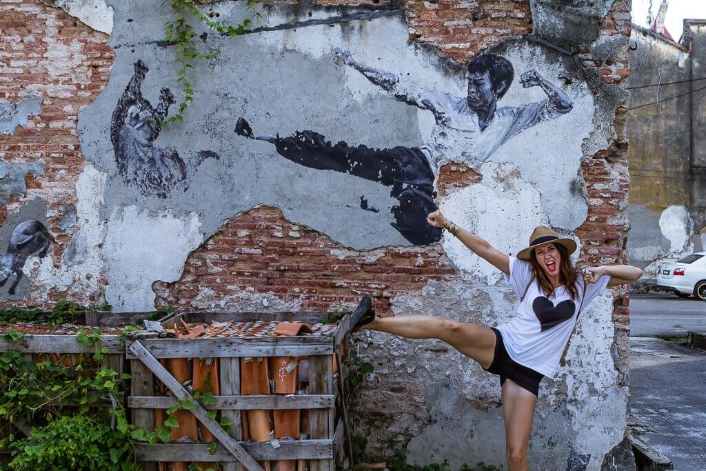 girl in hat and white t-shirt with black heart and black shorts pretending to kick at crate -- mimicking street art behind her of bruce lee in famous fighting pose on a gray and brick wall