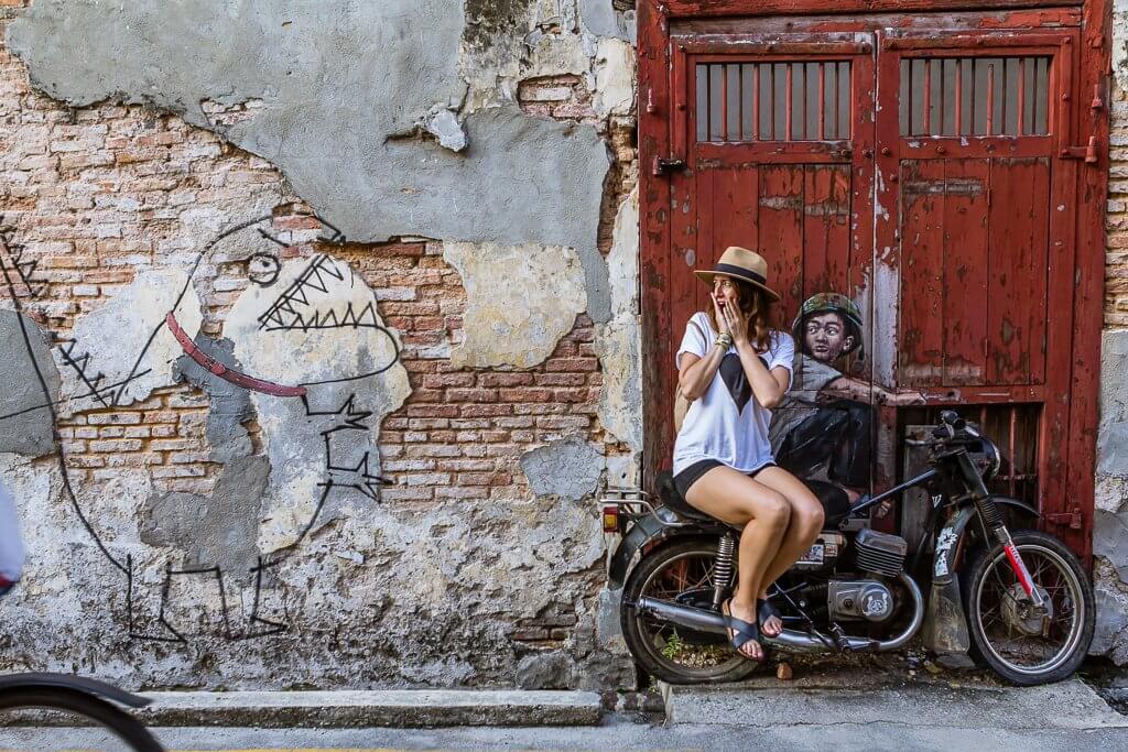 girl in straw hat, white t-shirt, and black shorts at street art scene in Penang -- street art is of a boy on a motorbike with a real motorbike in front on a red door to the right and a funny dinosaur outline to the left on a patchy brick wall.